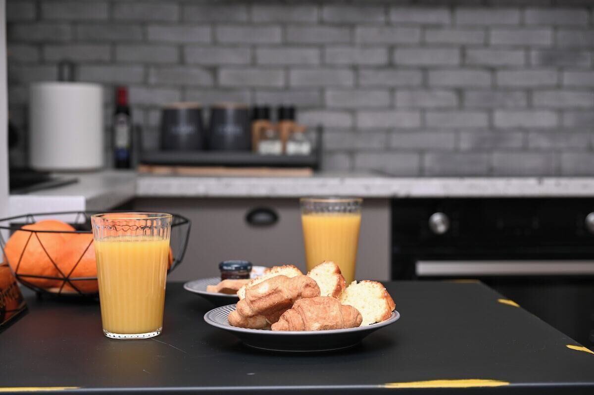 En frukostuppsättning med croissanter och apelsinjuice på ett bord, med en tegelvägg och köksredskap i bakgrunden.