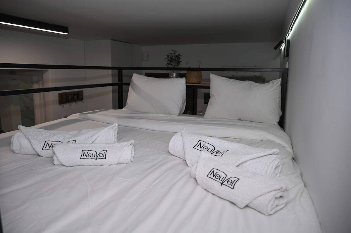 A neatly made bed with white linens and two towels with 'Neuvel' branding, a small plant on a shelf, and a 'HOME' sign on the wall.