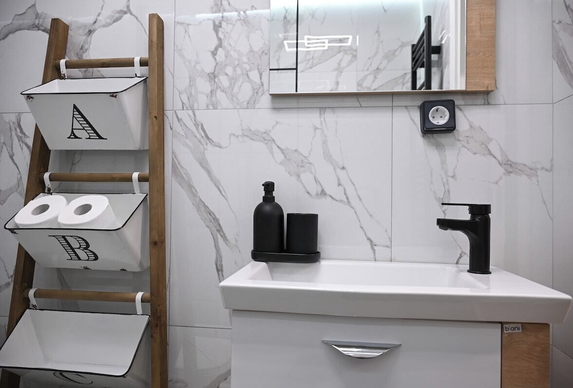 Een badkamer met een marmeren spatwand, een witte wastafel met een zwarte kraan, en een houten ladder met opbergkisten.