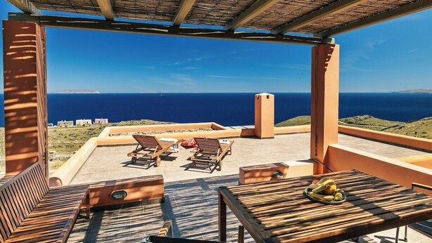 Un área de estar al aire libre con mobiliario de madera y una mesa con fruta, con vistas a la costa.