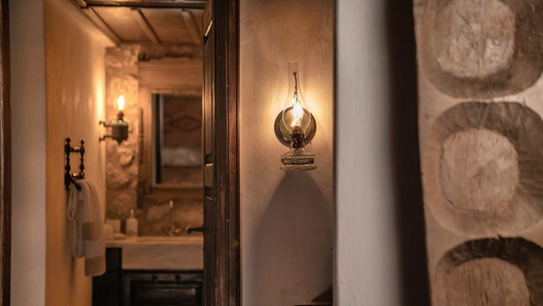 Un estrecho pasillo con una pared de piedra en un lado y un marco de puerta de madera en el otro. Se observa una lámpara fijada a la pared de piedra.