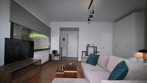 Un soggiorno moderno con una grande TV a schermo piatto, un divano beige con cuscini color acquamarina, un tavolino in legno e, sullo sfondo, una zona pranzo.