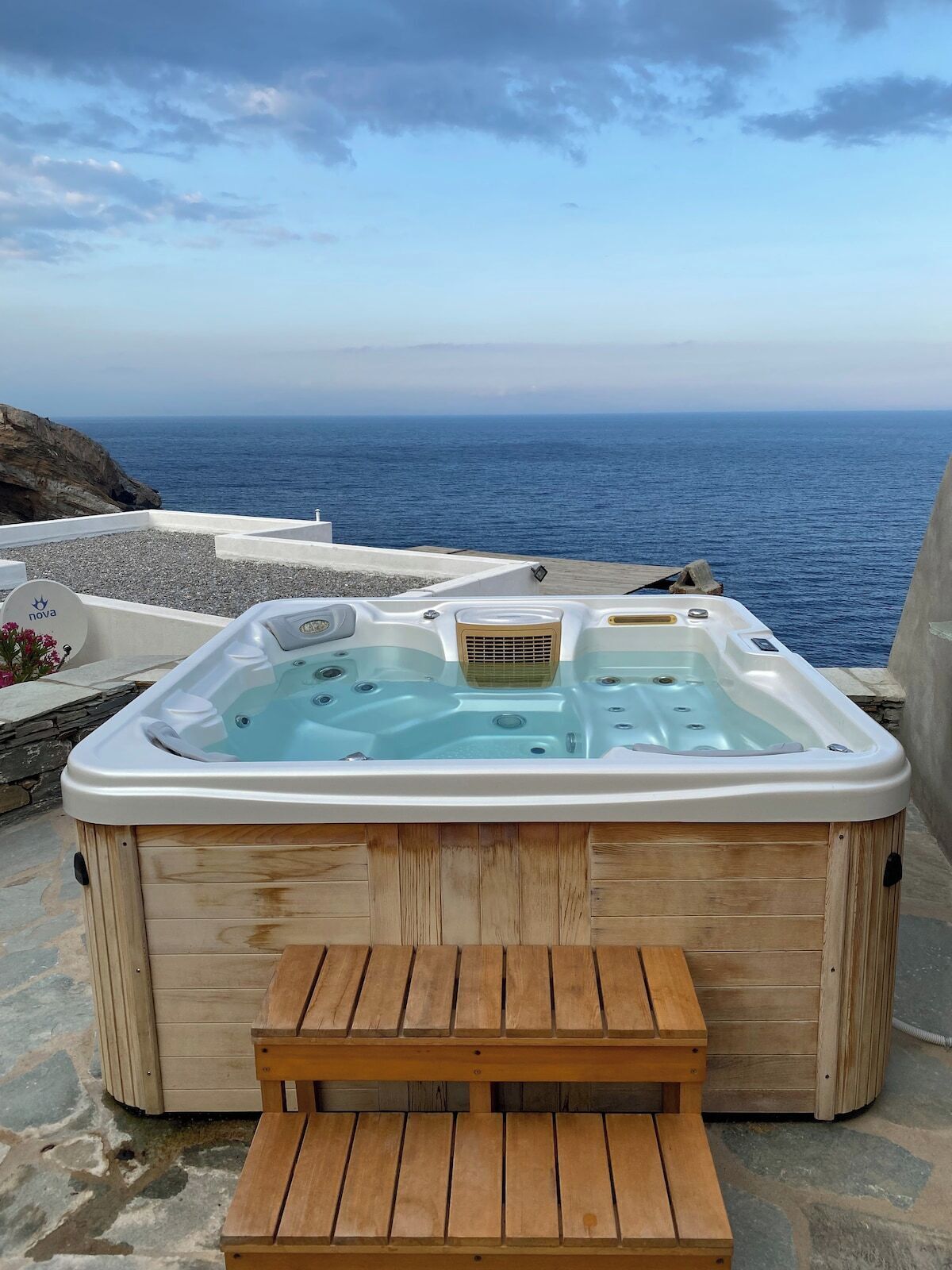 A wooden hot tub with a bench in front, set on a stone patio overlooking the sea.