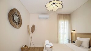A hotel room with a bed, a bedside table, a wall-mounted air conditioner, a decorative mirror, and a ceiling light fixture. - Coastal Charm by Neuvel (Glyfada)