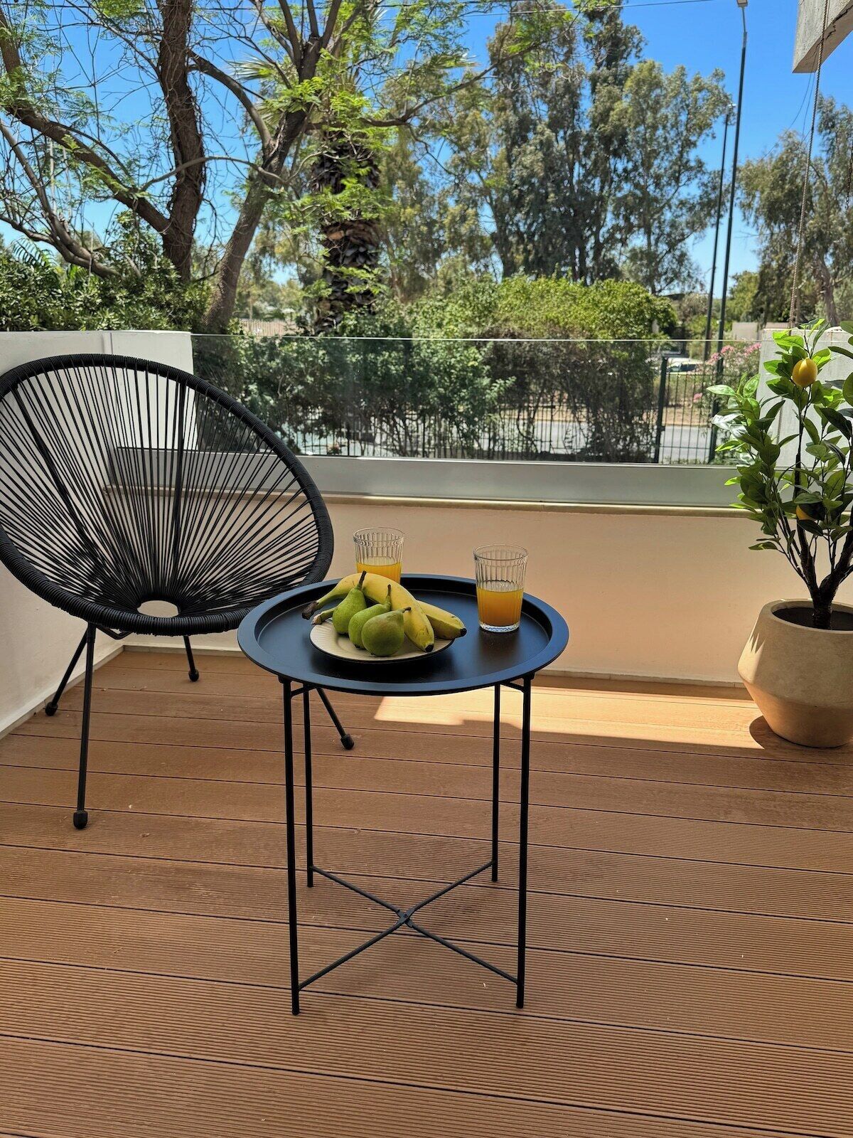 A wooden deck with a black chair, a small table with a plate of fruit and a glass of orange juice, and a potted plant.