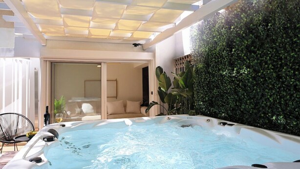 A hot tub area with a glass door leading to a seating area, surrounded by greenery and a pergola. - Coastal Charm by Neuvel (Glyfada)