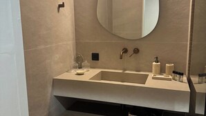 A modern bathroom with a beige tiled wall, a round mirror, a sink with a faucet, and a toilet. - Coastal Charm by Neuvel (Glyfada)