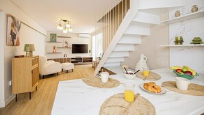 A modern living room with a staircase, a dining area with breakfast items, and a TV in the background. - Coastal Charm by Neuvel (Glyfada)