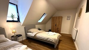 Quarto com teto inclinado, cama, mesa de cabeceira, guarda-roupa de madeira e janela com persianas.