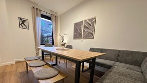 Uma área de refeições com uma mesa de madeira, quatro bancos, um sofá cinzento e dois painéis decorativos fixados na parede.
