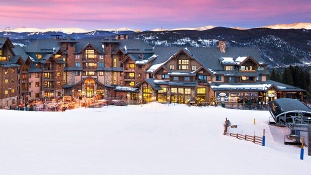 Un grande albergo con diversi edifici, una pista da sci in primo piano e montagne innevate sullo sfondo.