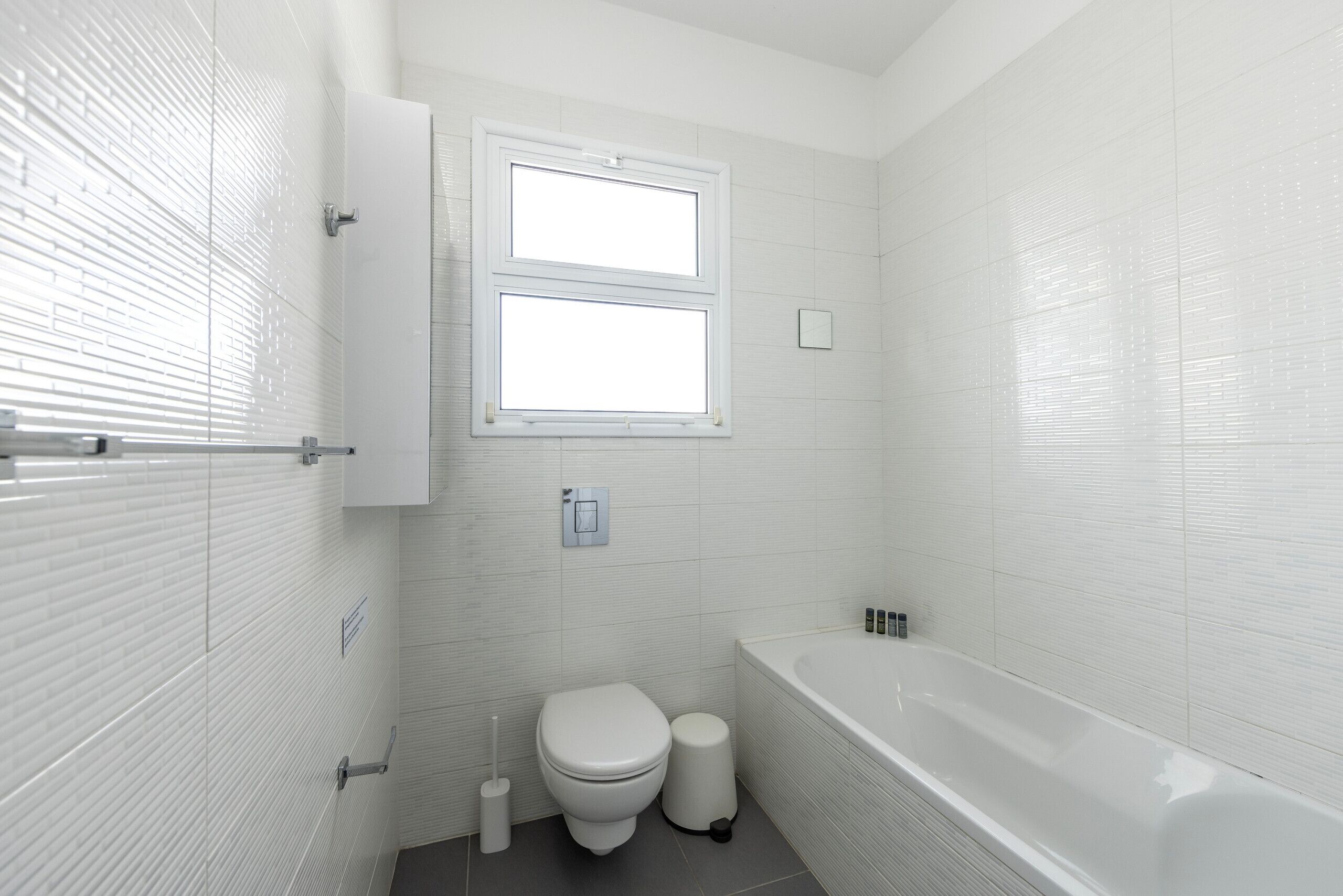 A modern bathroom with white tiles, a toilet, a bidet, a bathtub, and a window.