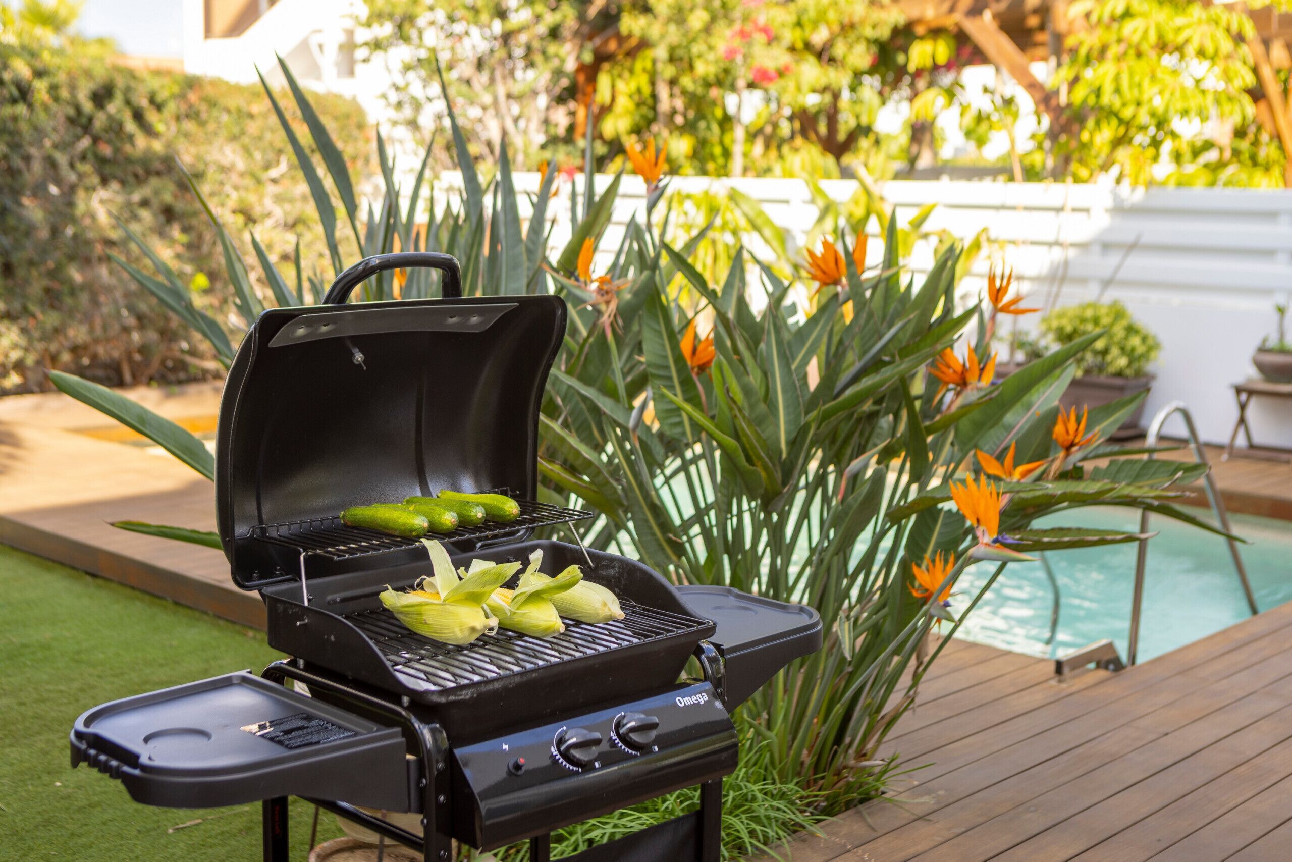 Un barbecue noir avec des épis de maïs, entouré de plantes et d’une piscine.