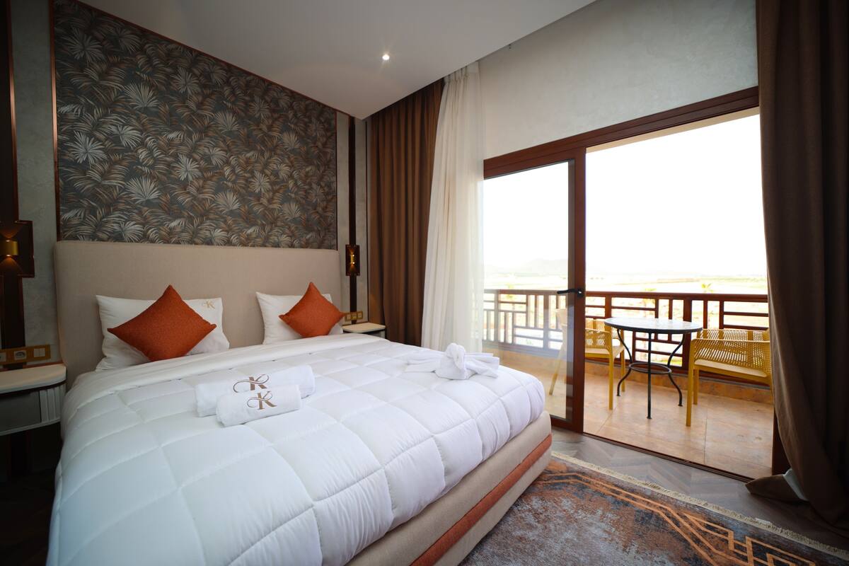 A hotel room with a large bed, a balcony with chairs, and a view of the landscape.