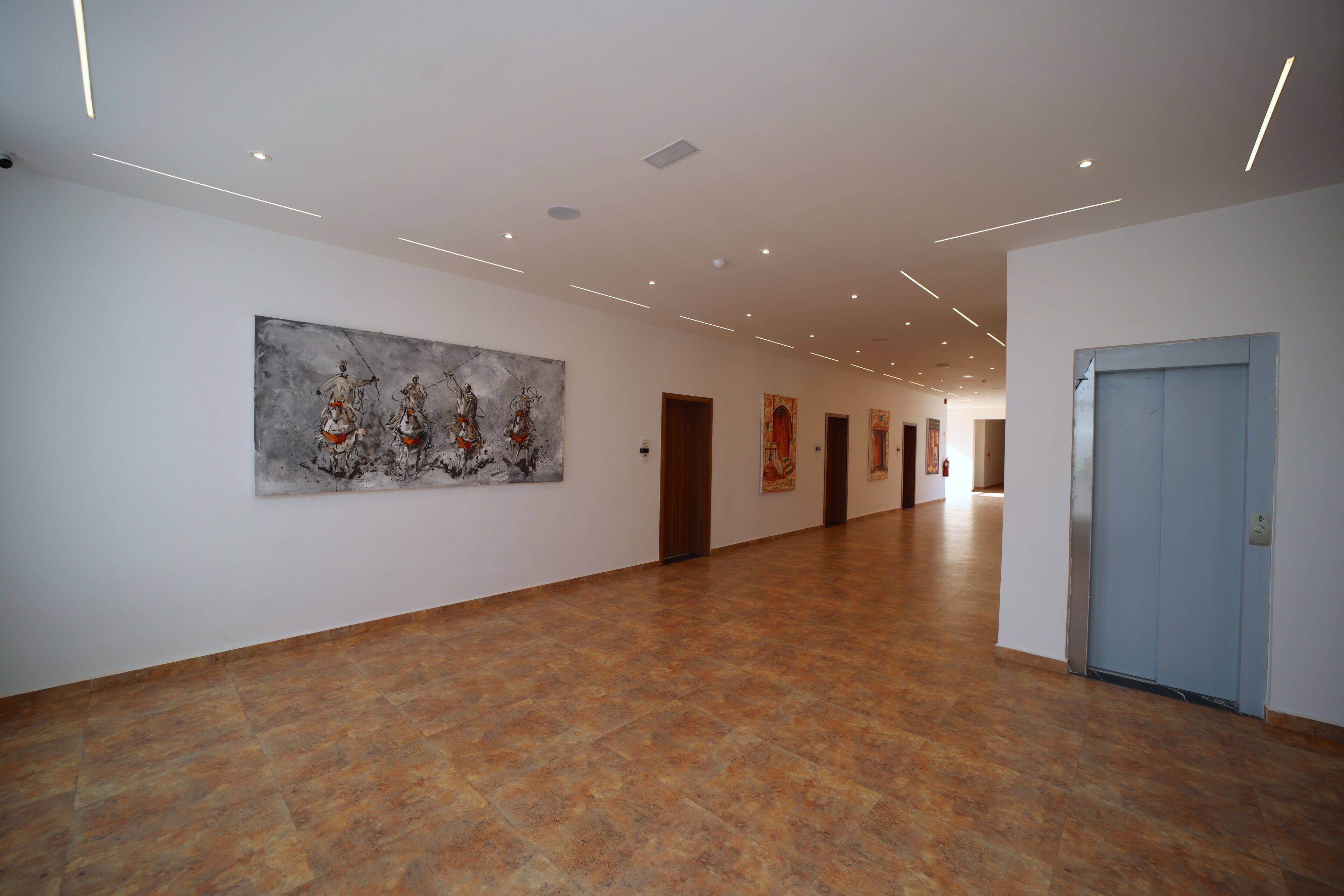 A spacious hallway with a large abstract painting on the left wall, a blue door on the right, and a tiled floor.