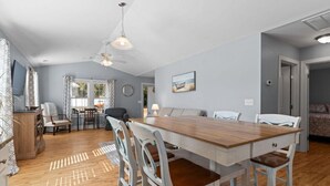 Un vaste espace dédié à la salle à manger, avec une table et des chaises en bois, un ventilateur de plafond et une vue sur l’extérieur grâce à de grandes fenêtres.