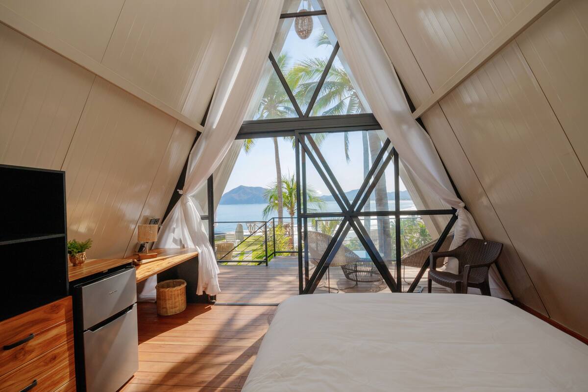Ein Schlafzimmer mit einem Bett, einem Schreibtisch und einem Stuhl. Ein Fenster mit Vorhängen bietet Blick auf Palmen und Berge.