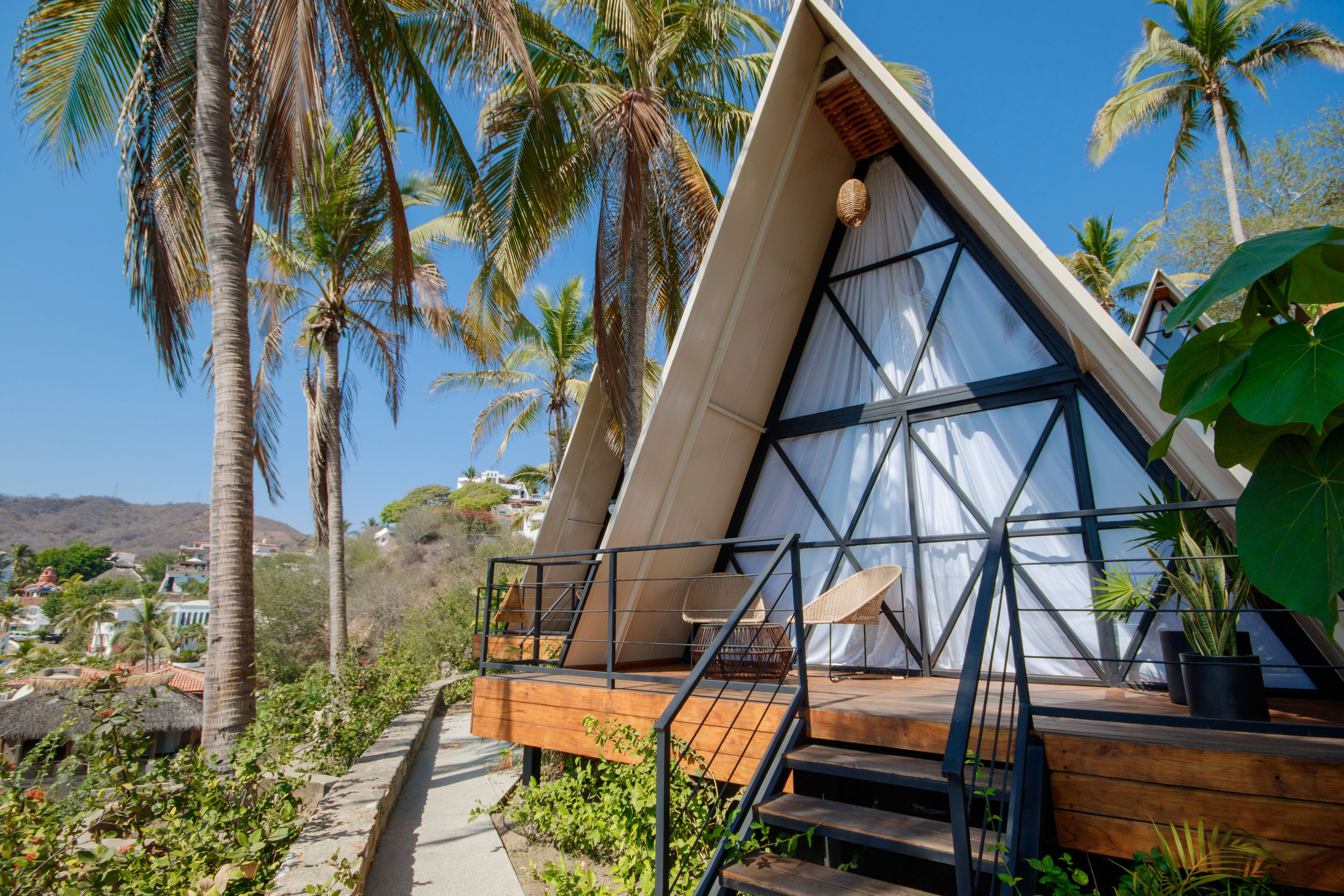 A modern, triangular-shaped building with a wooden deck and metal railings, surrounded by palm trees and greenery.