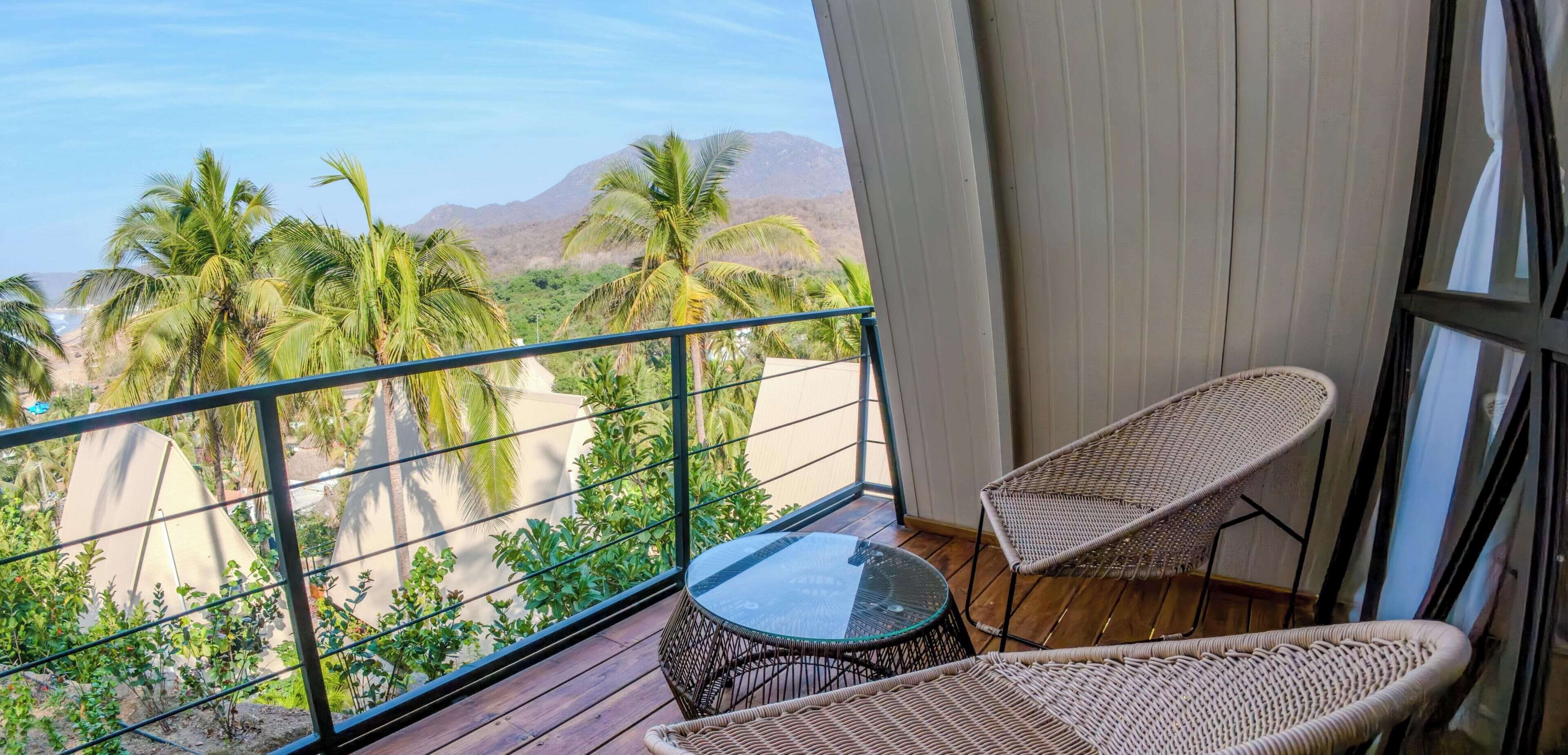 A balcony with a view of palm trees and mountains, wicker furniture, and a glass table.