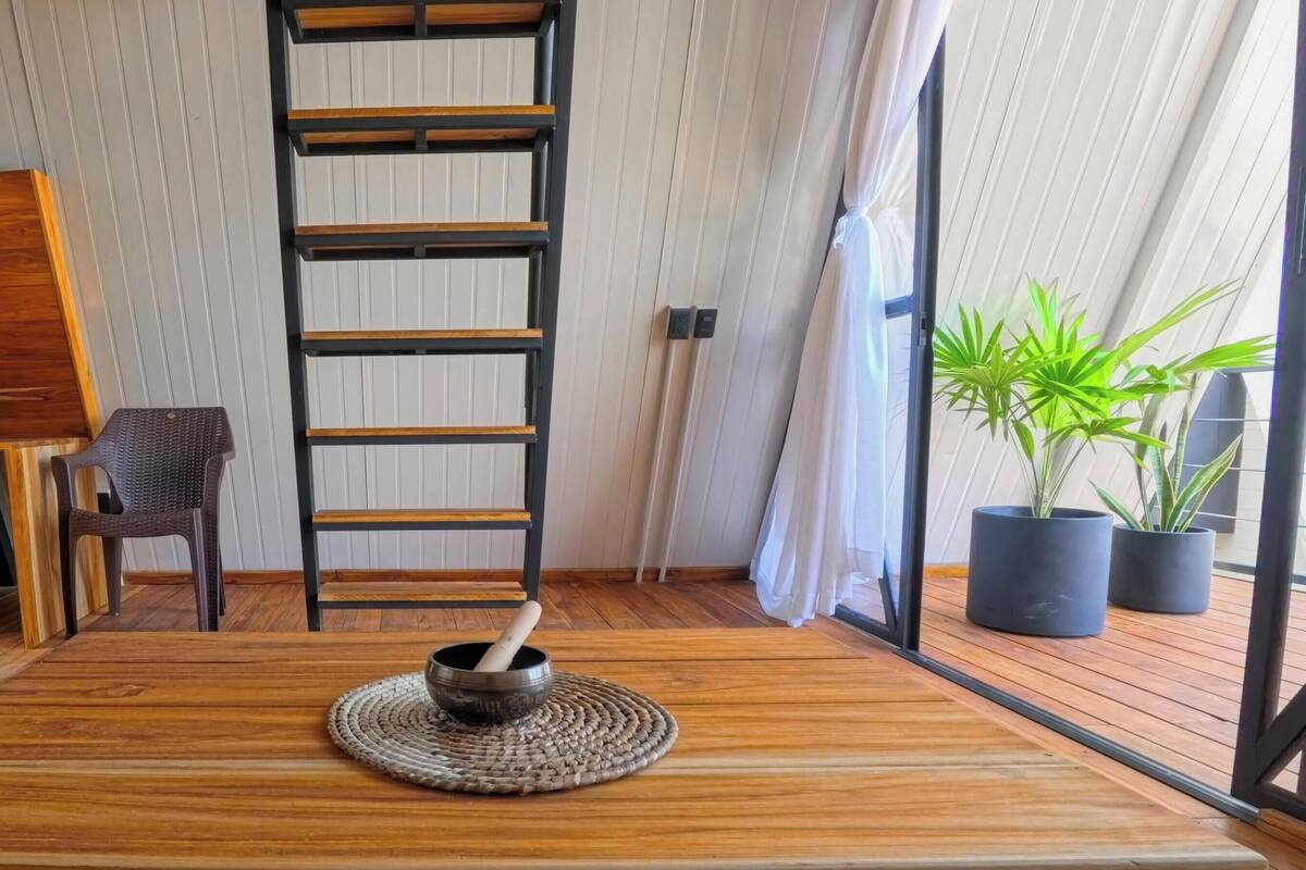 Ein Zimmer mit Holzboden, einem schwarzen Holzschrank, einem Korbstuhl, einer schwarzen Leiter, einem schwarzen Topf mit einem Holzhammer und Topfpflanzen.