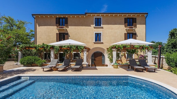 A poolside area with lounge chairs and umbrellas, a clear blue pool, and a building with a tiled roof and arched entrance. - Villa Christina by Interhome (Tinjan)