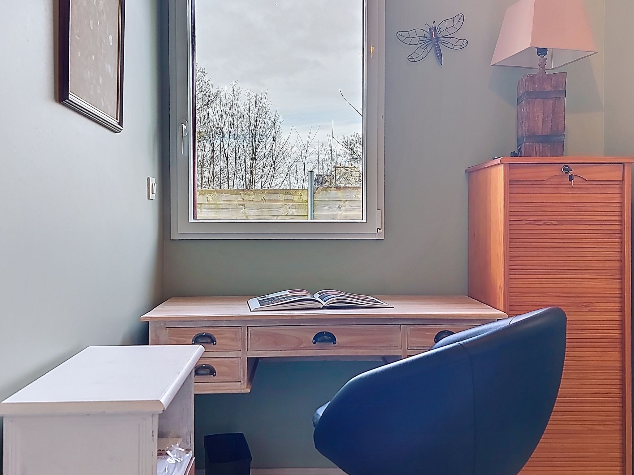 A room with a desk, chair, and a window with a view of trees and a fence.