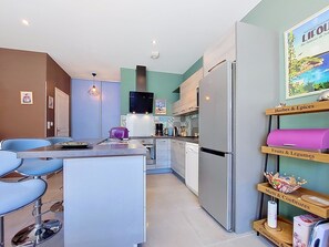 Eine moderne Küche mit einer zentralen Kochinsel, Barhocker, einem Kühlschrank und einem Regal mit beschrifteten Fächern für Kräuter, Früchte und Minze.