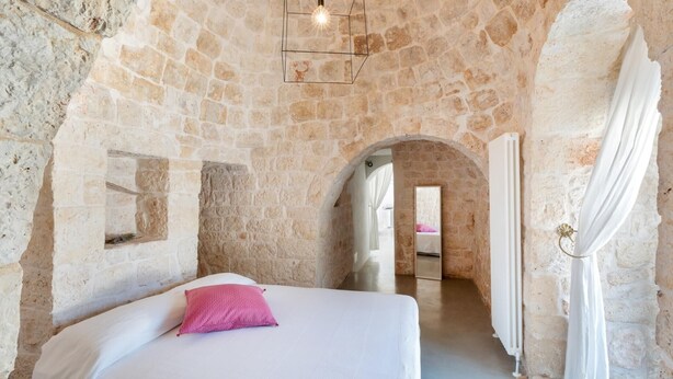 Une pièce aux murs de pierre, avec un lit, un luminaire suspendu et une porte en arc de cercle.