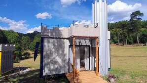 Una pequeña estructura de estilo castillo con una terraza de madera y una sola puerta, rodeada de césped y árboles.