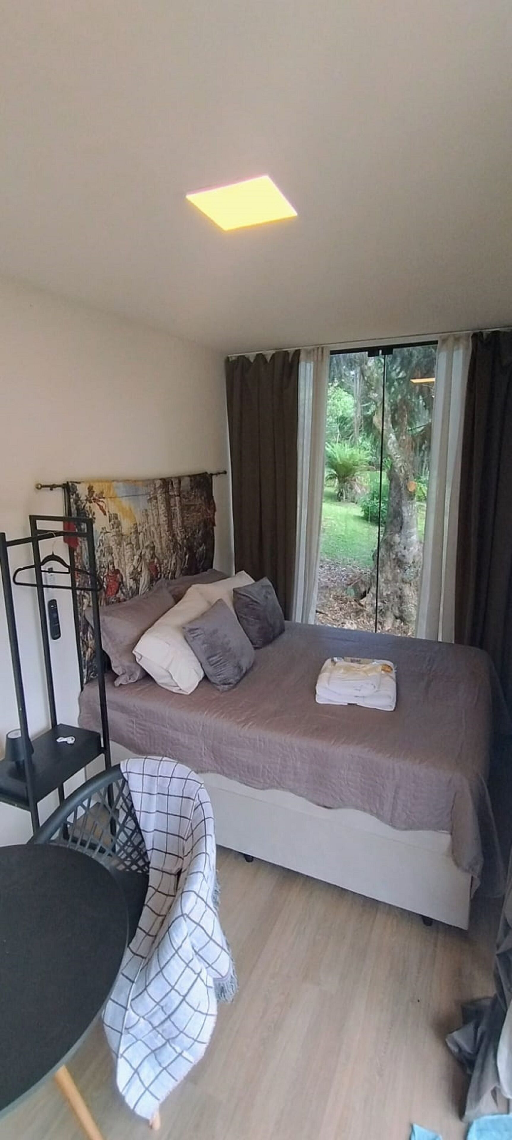 Un dormitorio con cama, una silla, una mesa, perchero y ventana con cortinas.