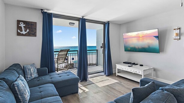 A living room with a large sofa, a coffee table, and a painting of a sunset. There is a sliding glass door leading to a balcony with a view of the ocean. - 🌞Gulf Views⛱️Outdoor Pool | Sauna | Walk to Beach | SunDestin 405☀️ (Destin)
