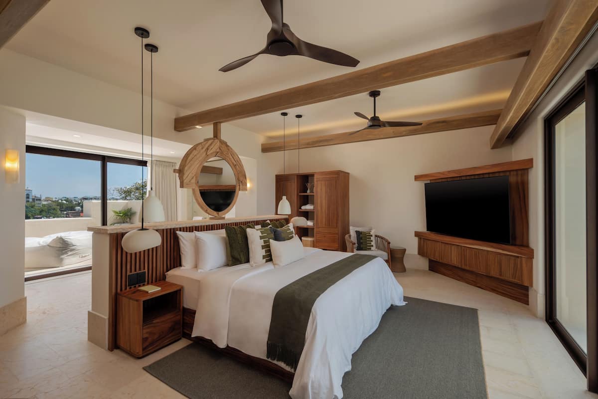 A modern bedroom with a large bed, wooden beams, a ceiling fan, a TV, and a view of the city.