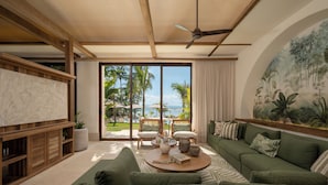 Ein großzügiger Wohnbereich mit einem großen Fernseher, grünen Sofas, einem Couchtisch und Blick auf Palmen und das Meer.