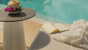 Ein Poolbereich mit Liegestuhl, Tisch mit Getränk und Blumen sowie Blick auf Strand und Meer.
