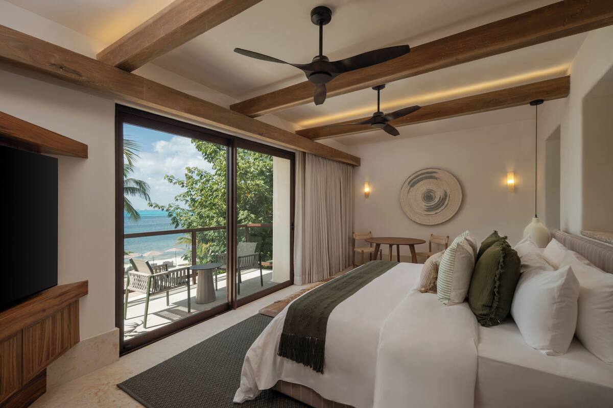A bedroom with a bed, a TV, a ceiling fan, a view of the ocean, and a balcony with outdoor seating.