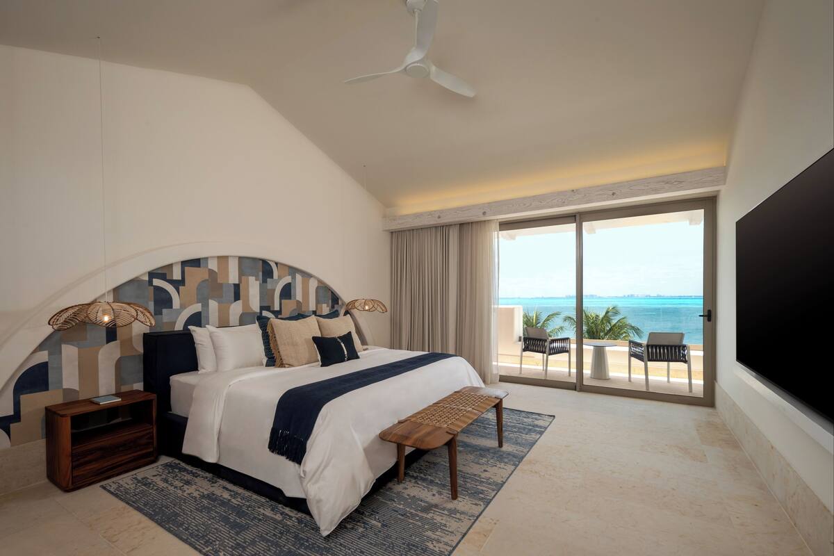 A bedroom with a large bed, a ceiling fan, a TV, and a balcony with a view of the ocean.