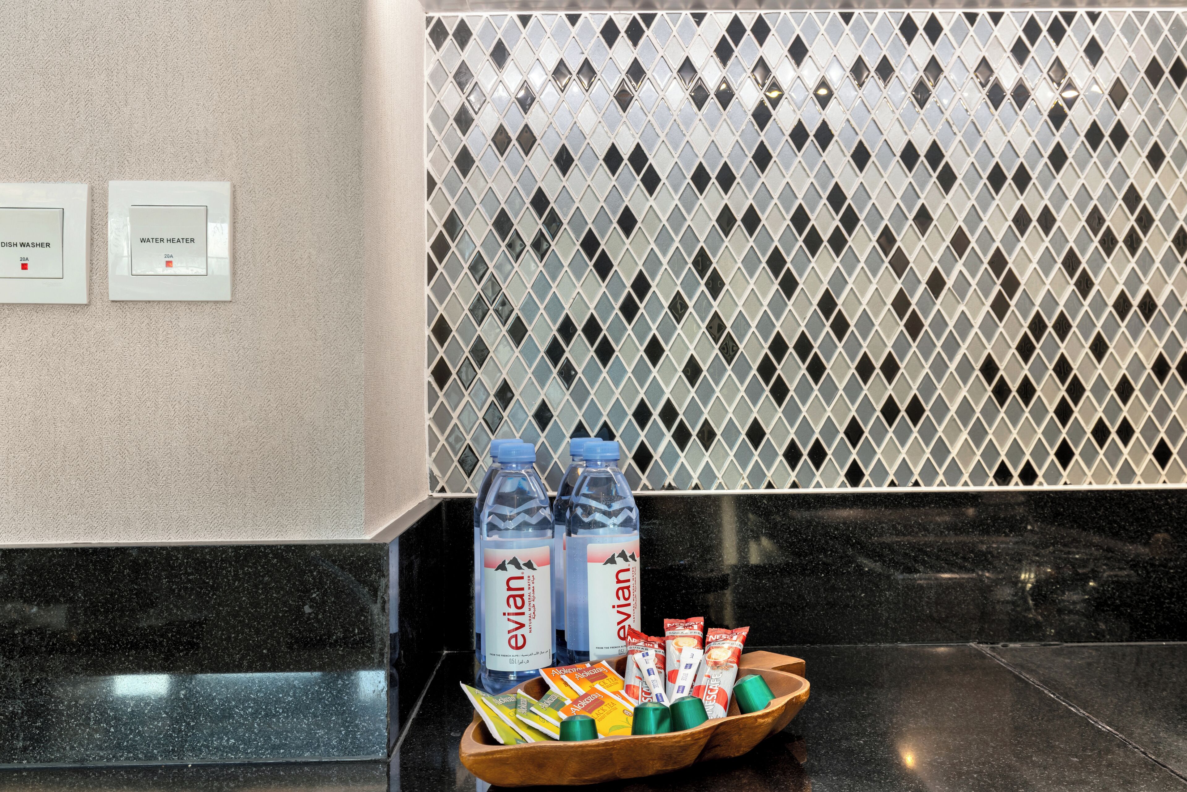 Zona de recepción de un hotel con mostrador de mármol negro, botellas de agua Evian y una bandeja con aperitivos.