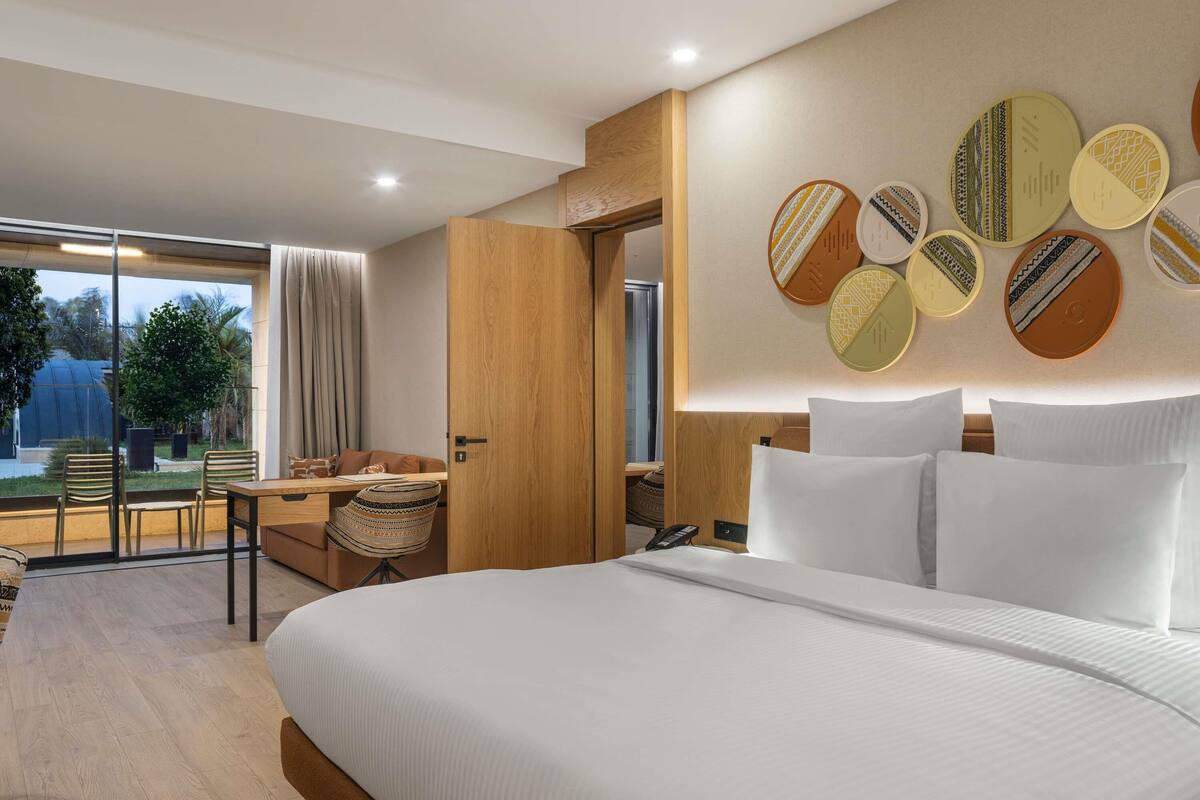 A modern hotel room with a large bed, a desk with chairs, a view through a glass door, and a wall with patterned decor.