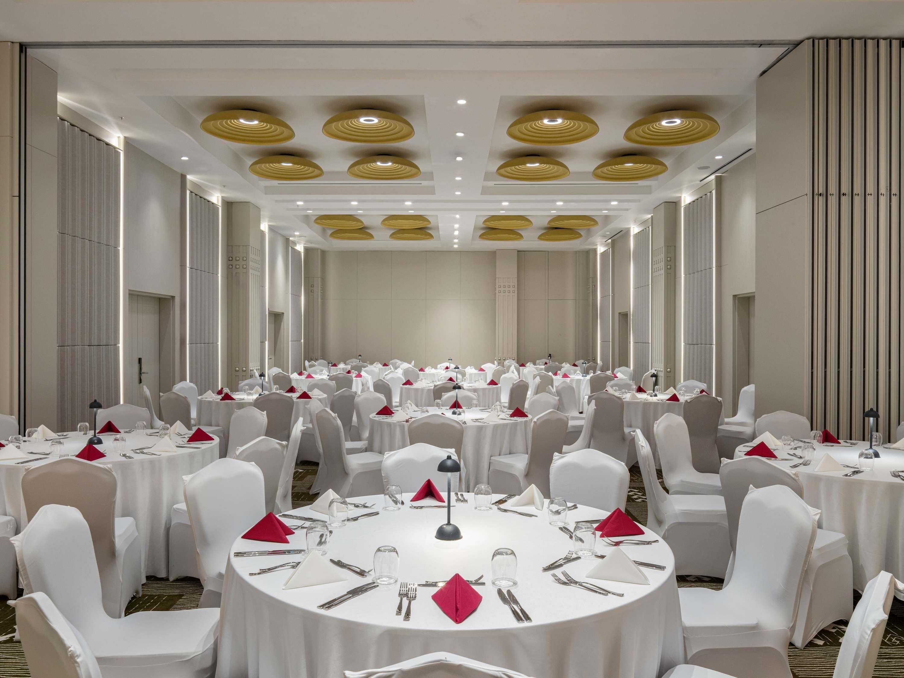 A large conference room with multiple round tables, white chairs, and red napkins.