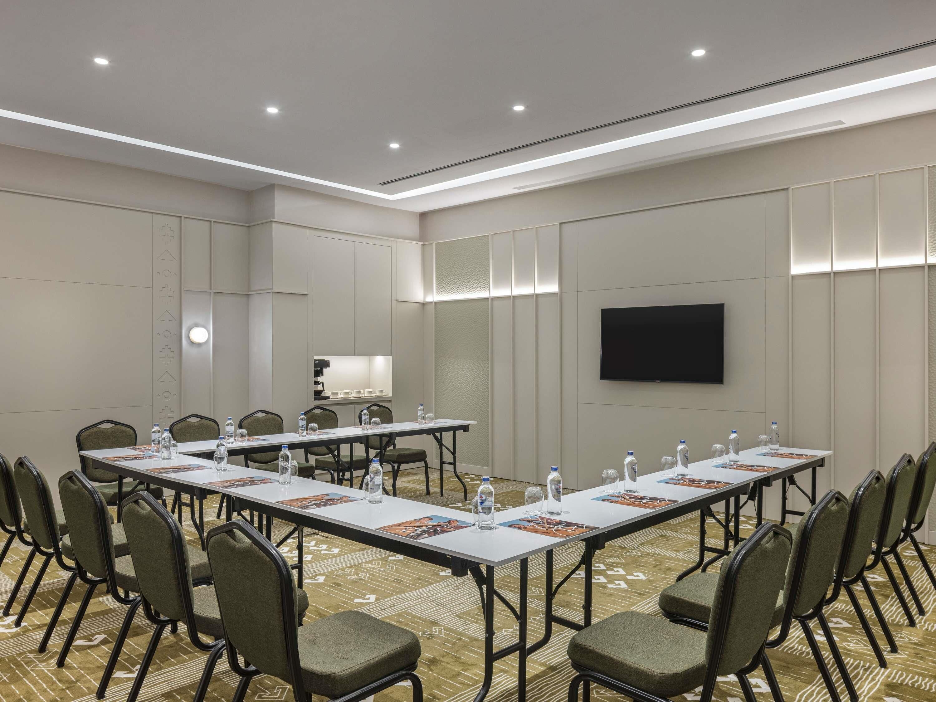 A conference room with long tables, chairs, water bottles, and a flat-screen TV.