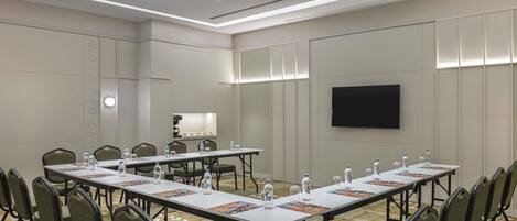 A conference room with long tables, chairs, water bottles, and a flat-screen TV.