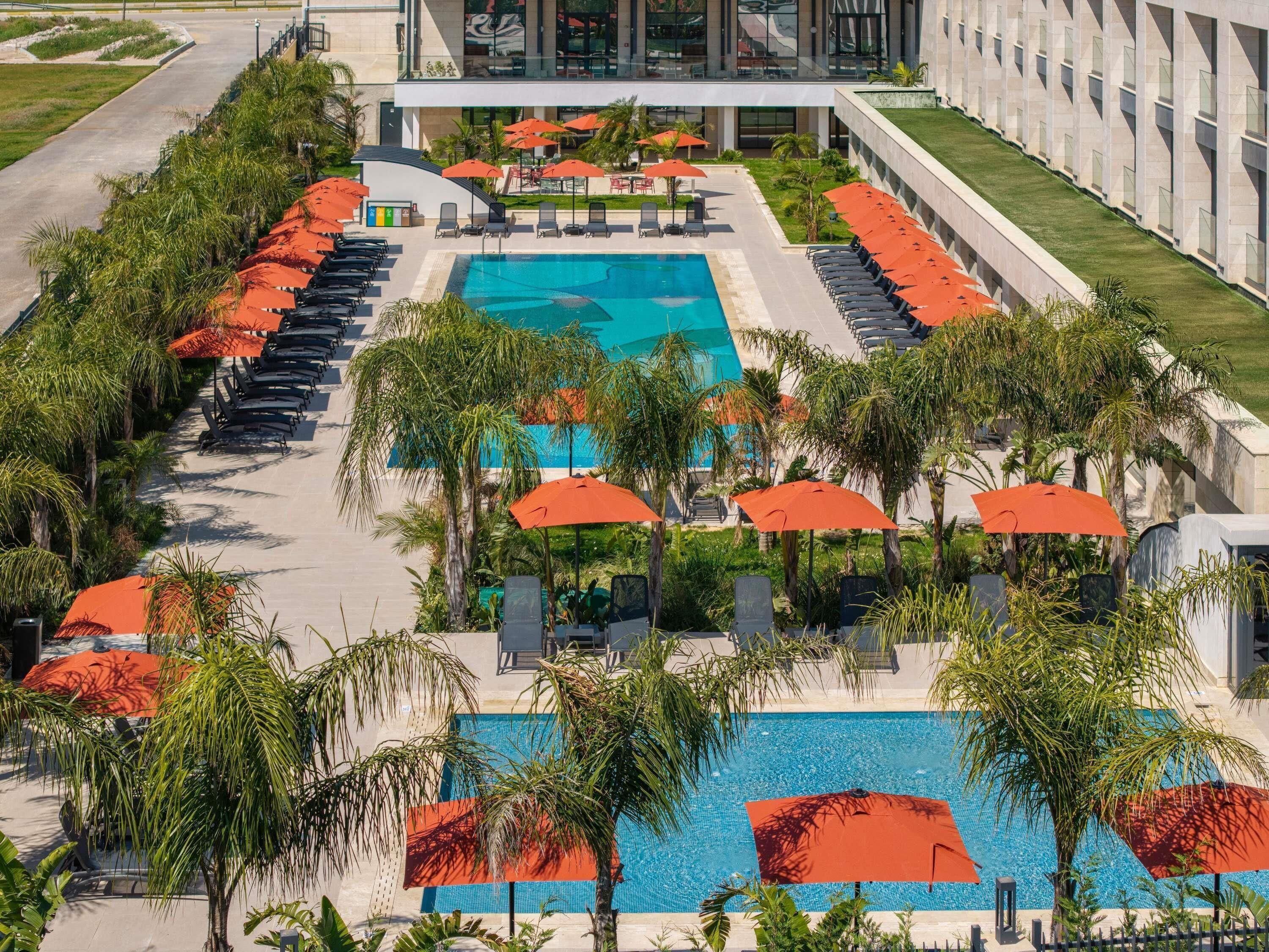 A large swimming pool surrounded by lounge chairs and umbrellas, with a modern building in the background.
