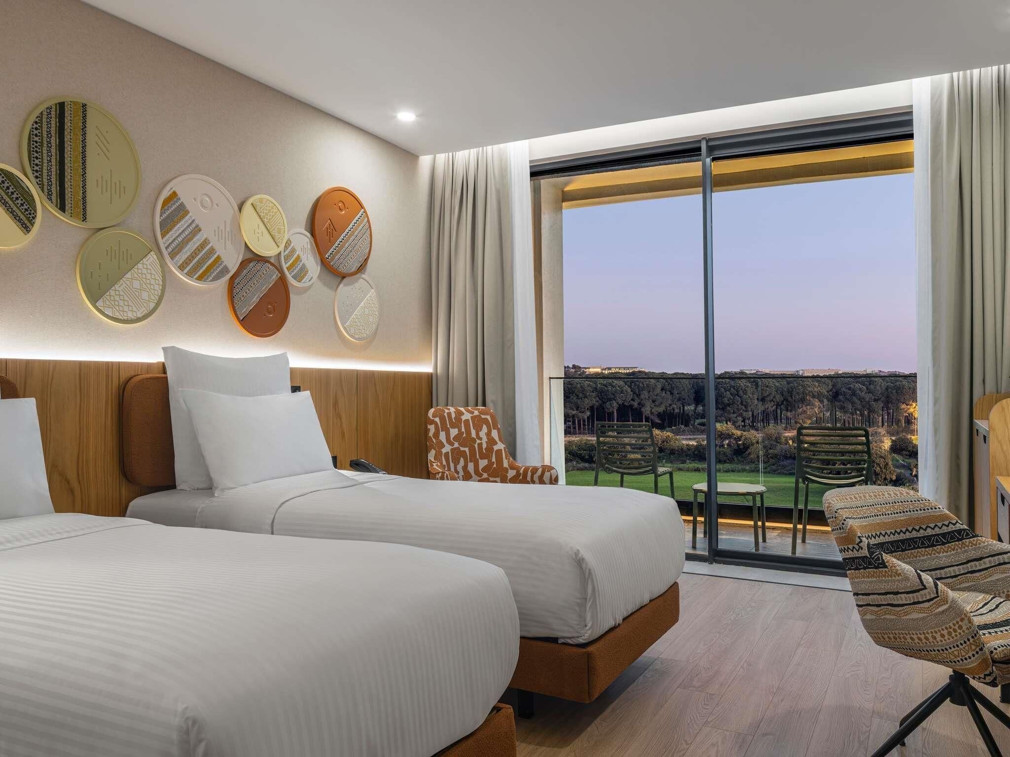 A hotel room with two beds, a large window with a city view, and a dining area with a table and chairs.