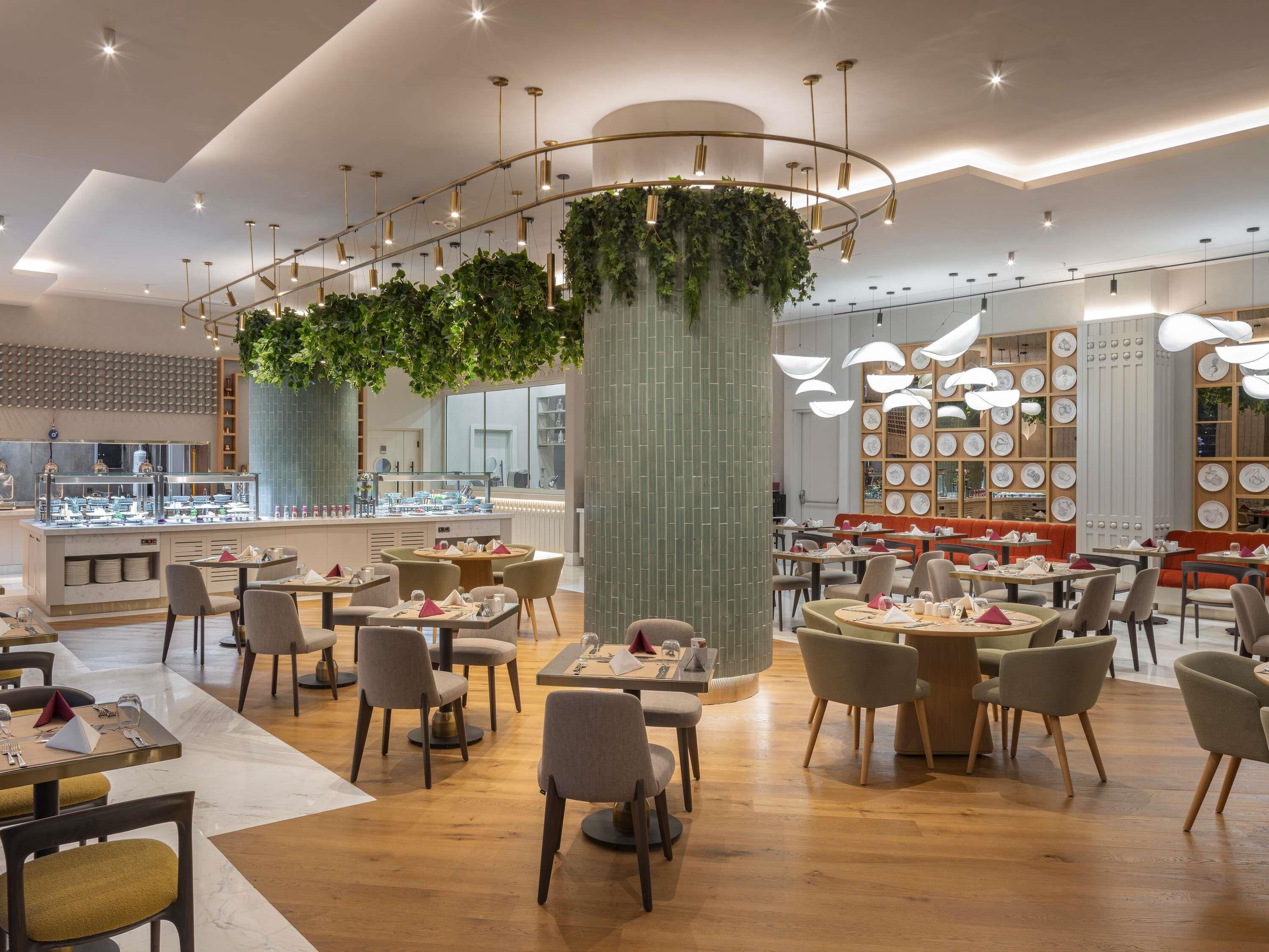 A modern restaurant interior with round tables, chairs, a central column with greenery, and a wall of plates.