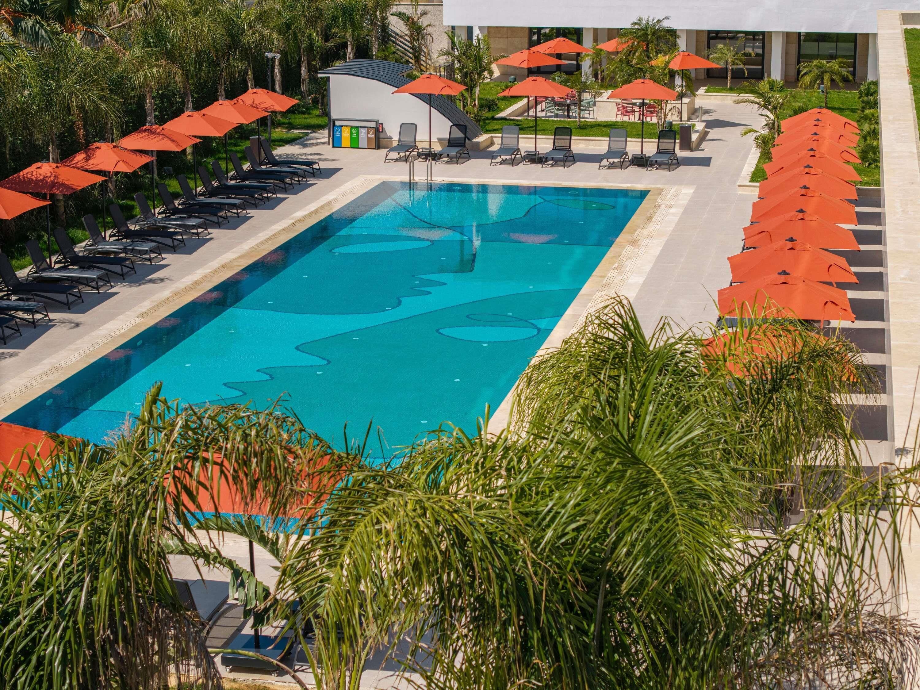 A swimming pool with lounge chairs and umbrellas, surrounded by palm trees and a building in the background.
