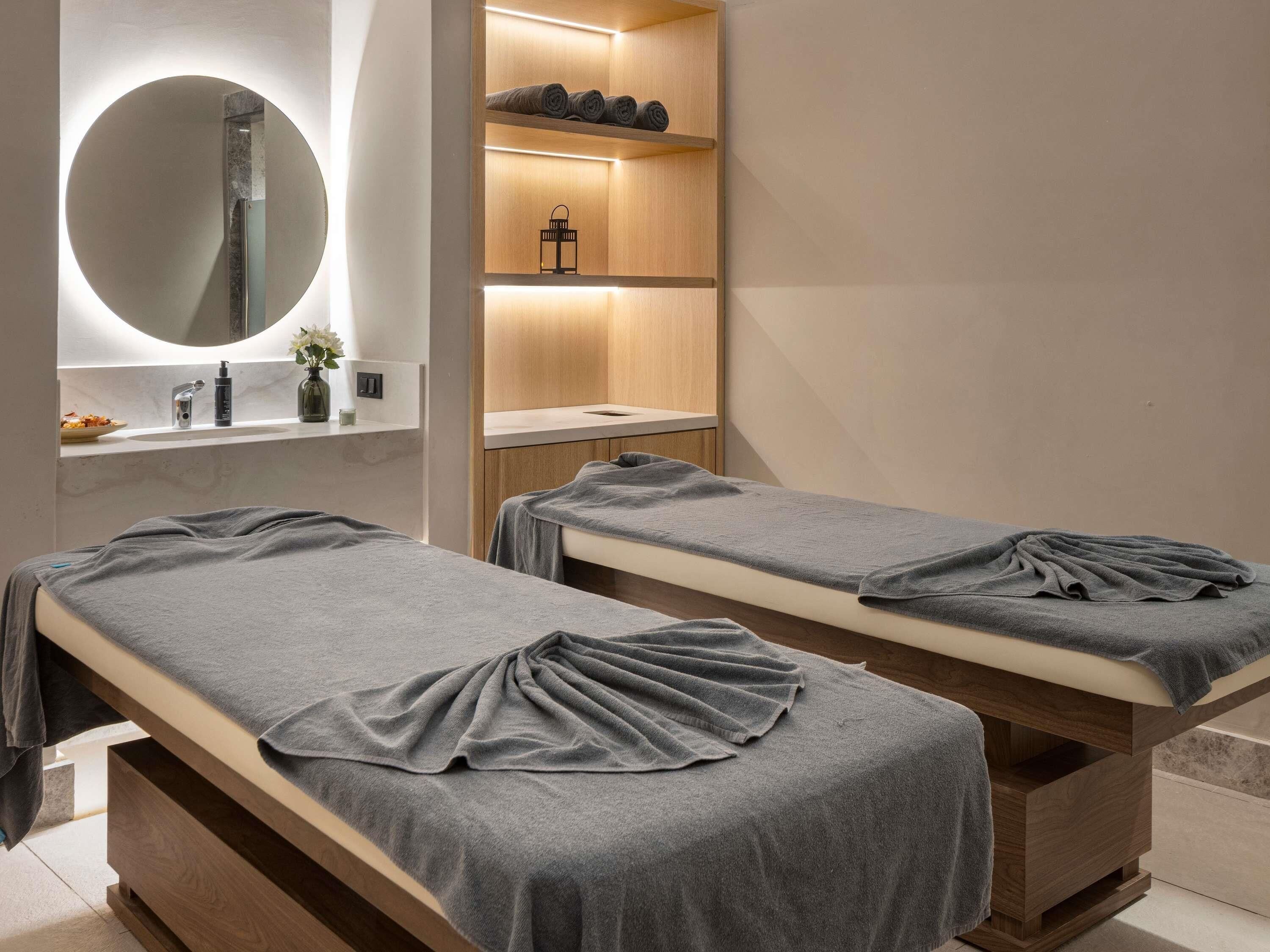 Un spa moderno con dos camillas de tratamiento, un tocador de madera con espejo y estanterías con toallas y un farol.