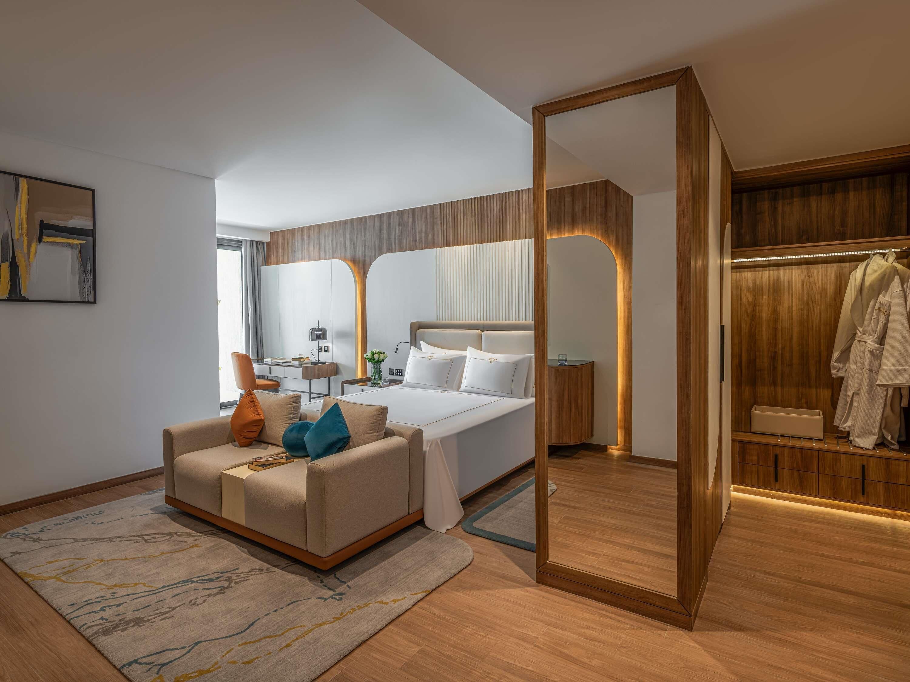 A modern hotel room with a bed, a seating area, a desk, a mirror, and a wardrobe.