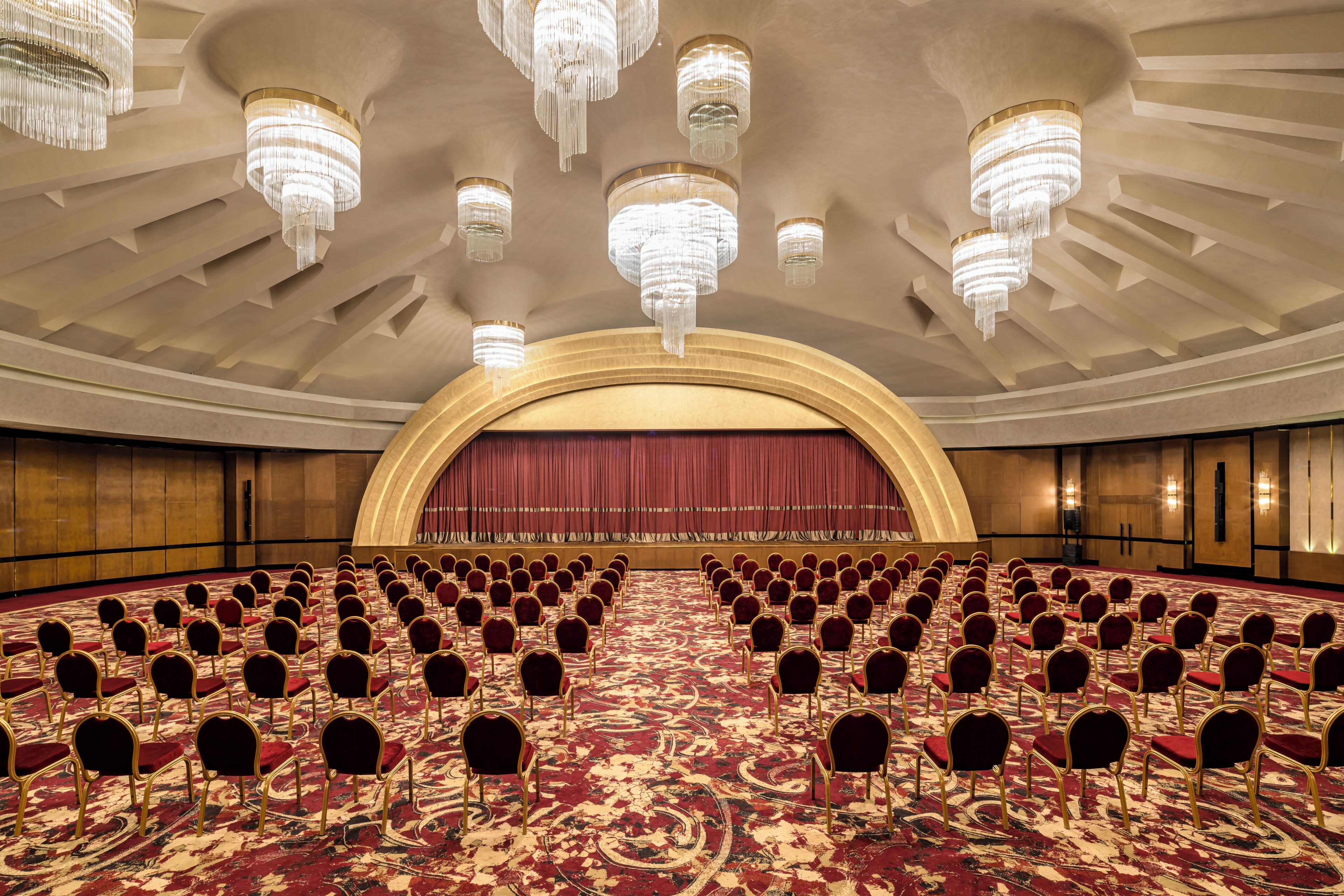 Un amplio salón de conferencias con filas de sillas, un escenario con cortina roja y varios candelabros.