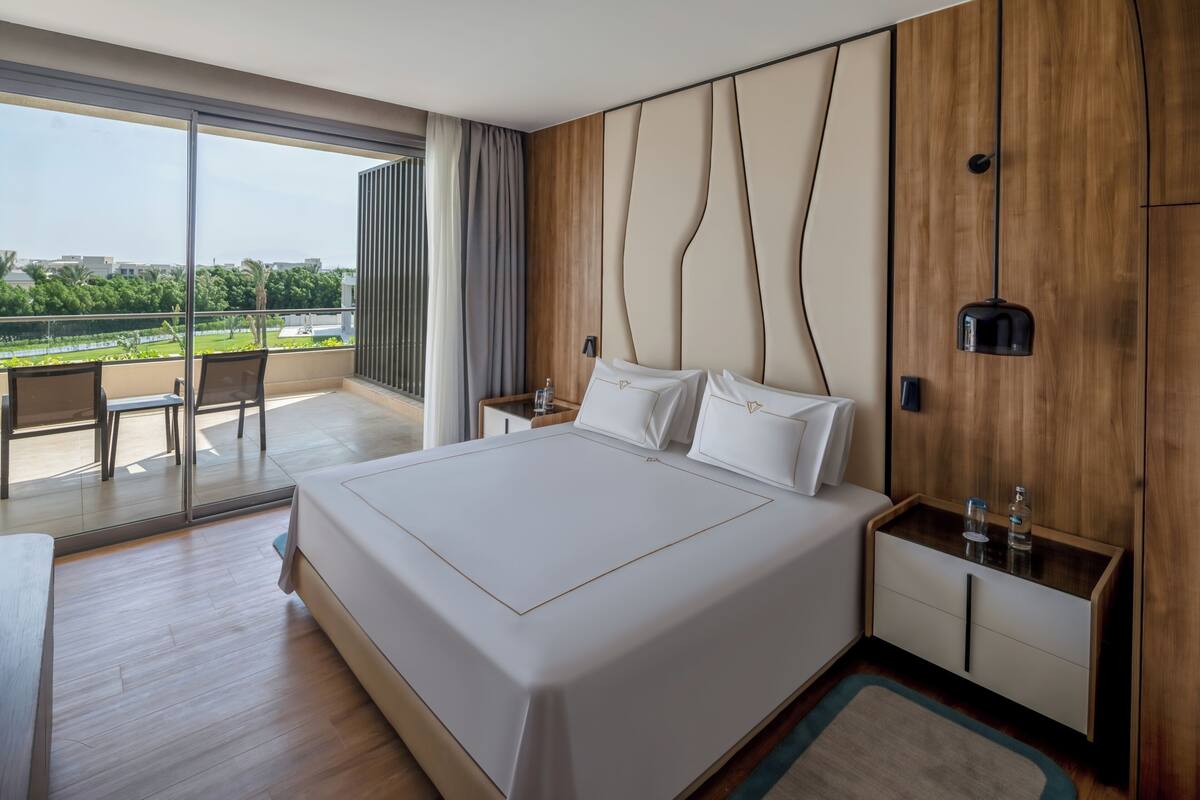 Une chambre d’hôtel moderne avec un grand lit, une tête de lit en bois, une table de chevet et une vue sur l’extérieur.