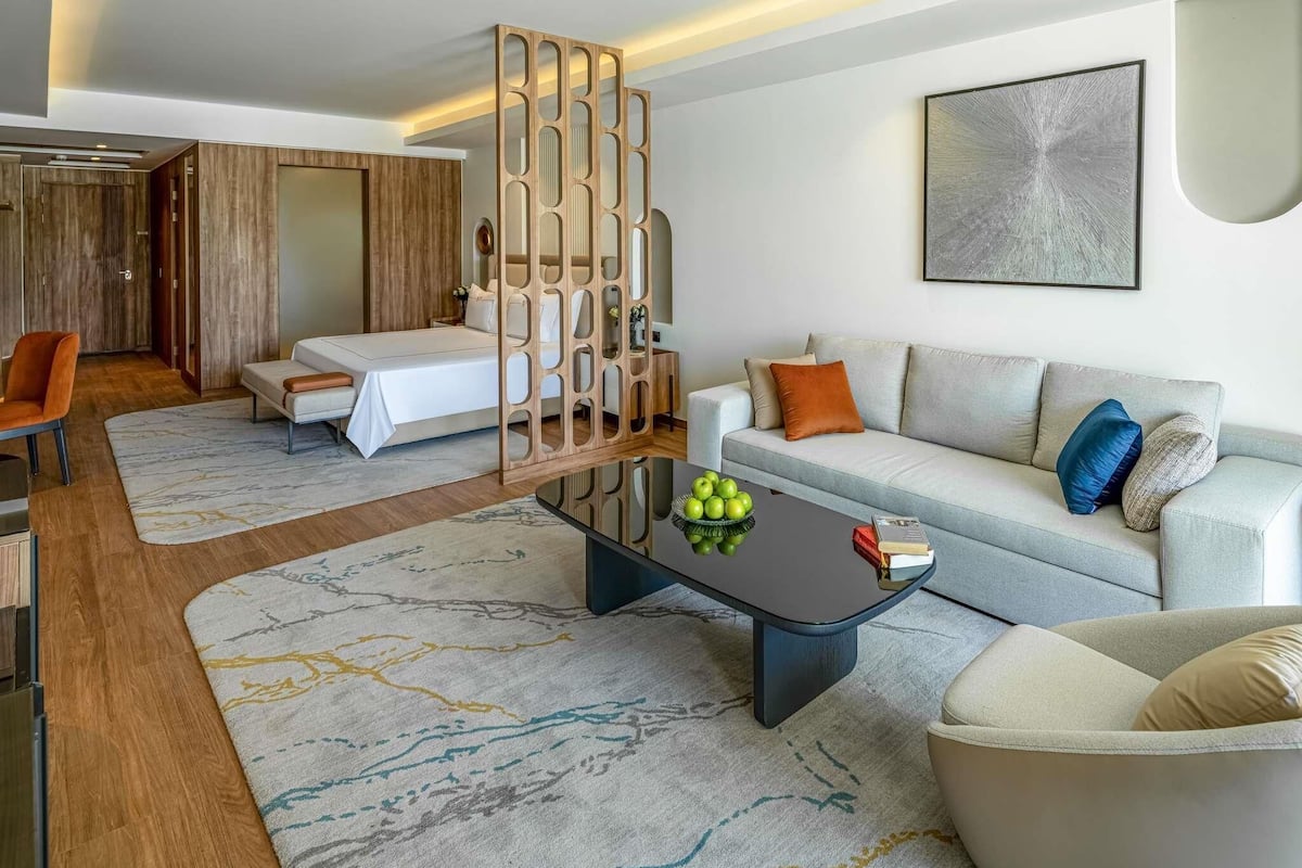 A modern hotel room with a bed, a sofa, a coffee table with fruit, a decorative screen, and a painting on the wall.